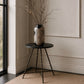 A decorative ceramic vase with a black and beige geometric pattern sits on a small round black side table in a styled interior corner with a window, beige wall molding, and a textured rug.