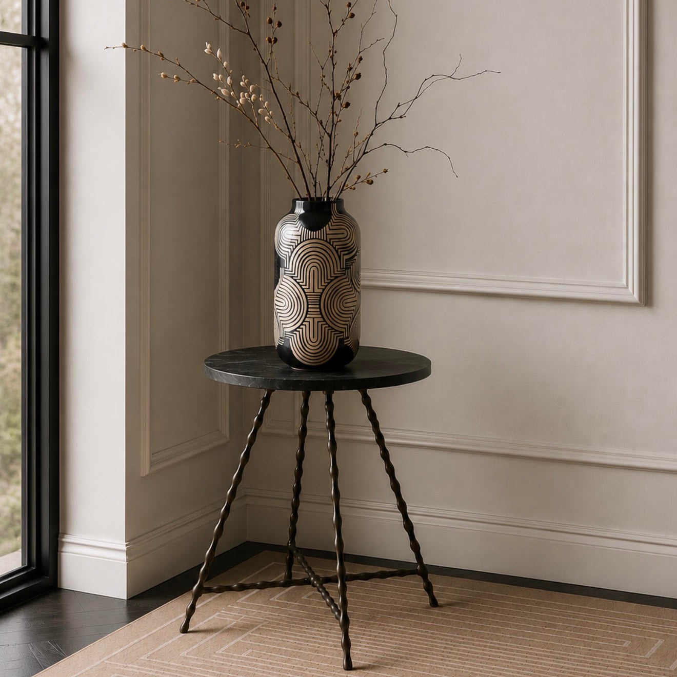 A decorative ceramic vase with a black and beige geometric pattern sits on a small round black side table in a styled interior corner with a window, beige wall molding, and a textured rug.