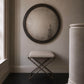 Round wall mirror with a dark wood frame hanging above a small upholstered stool with a dark metal frame, set in a dimly lit room with dark wood floors and a light wall.
