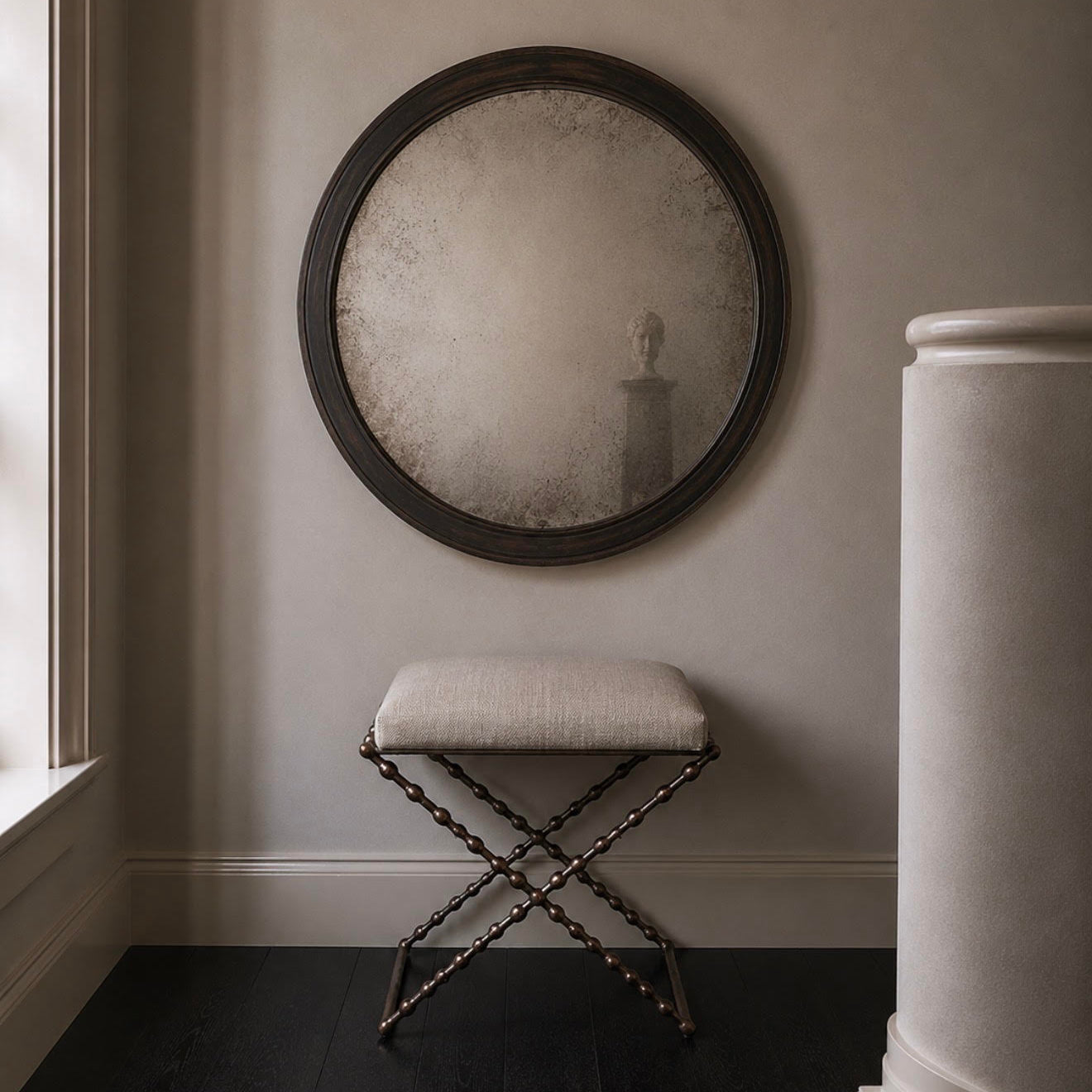 Round wall mirror with a dark wood frame hanging above a small upholstered stool with a dark metal frame, set in a dimly lit room with dark wood floors and a light wall.