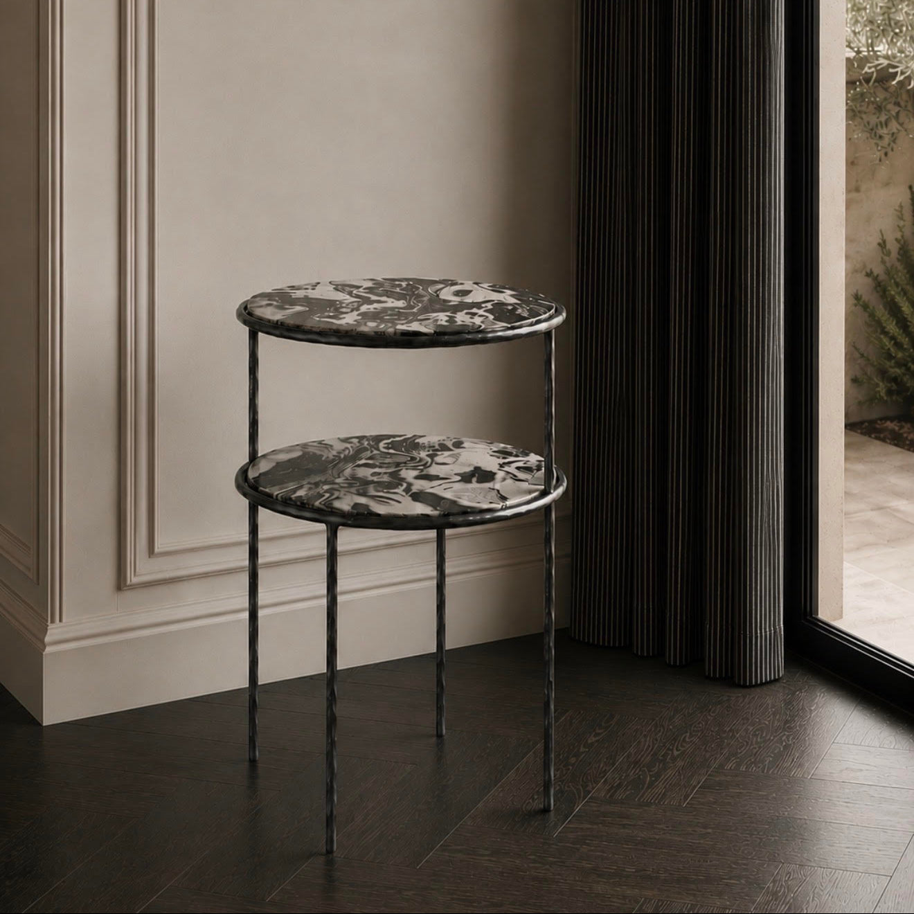 A two-tier round nesting side table with black marble-patterned tops and twisted dark metal legs, set in a modern interior next to a window with dark curtains.