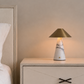 A modern table lamp with a white marble base and a brushed gold conical shade, glowing with a warm light on a cream-colored nightstand next to a bed.