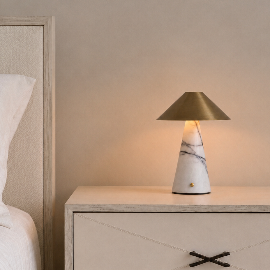 A modern table lamp with a white marble base and a brushed gold conical shade, glowing with a warm light on a cream-colored nightstand next to a bed.