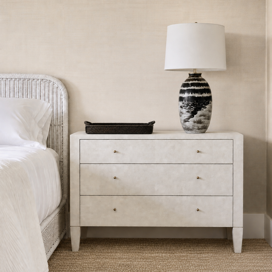 A three-drawer nightstand in a light textured finish stands beside a bed, styled with a black tray and a black-and-white patterned table lamp on top, against a beige wall and woven rug.
