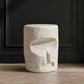 A cylindrical white stone stool with a textured, speckled surface and an abstract carved recess on the front, set against a dark paneled wall and light wood floor.