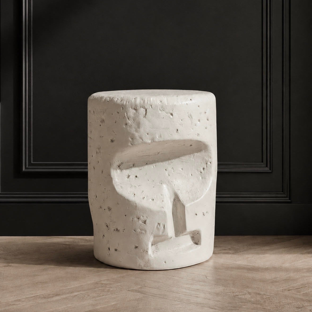 A cylindrical white stone stool with a textured, speckled surface and an abstract carved recess on the front, set against a dark paneled wall and light wood floor.
