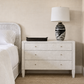 A three-drawer nightstand in a light textured finish stands beside a bed, styled with a black tray and a black-and-white patterned table lamp on top, against a beige wall and woven rug.