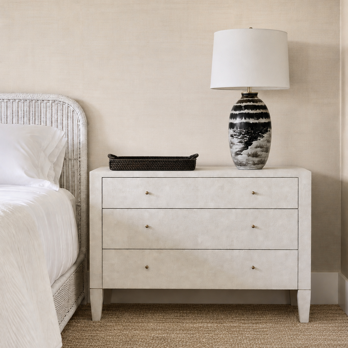 A three-drawer nightstand in a light textured finish stands beside a bed, styled with a black tray and a black-and-white patterned table lamp on top, against a beige wall and woven rug.