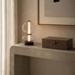 A modern table lamp with a clear glass double-sphere design and a vertical illuminated light bar, placed on a textured stone console table next to a dark wood box in a minimalist interior setting.