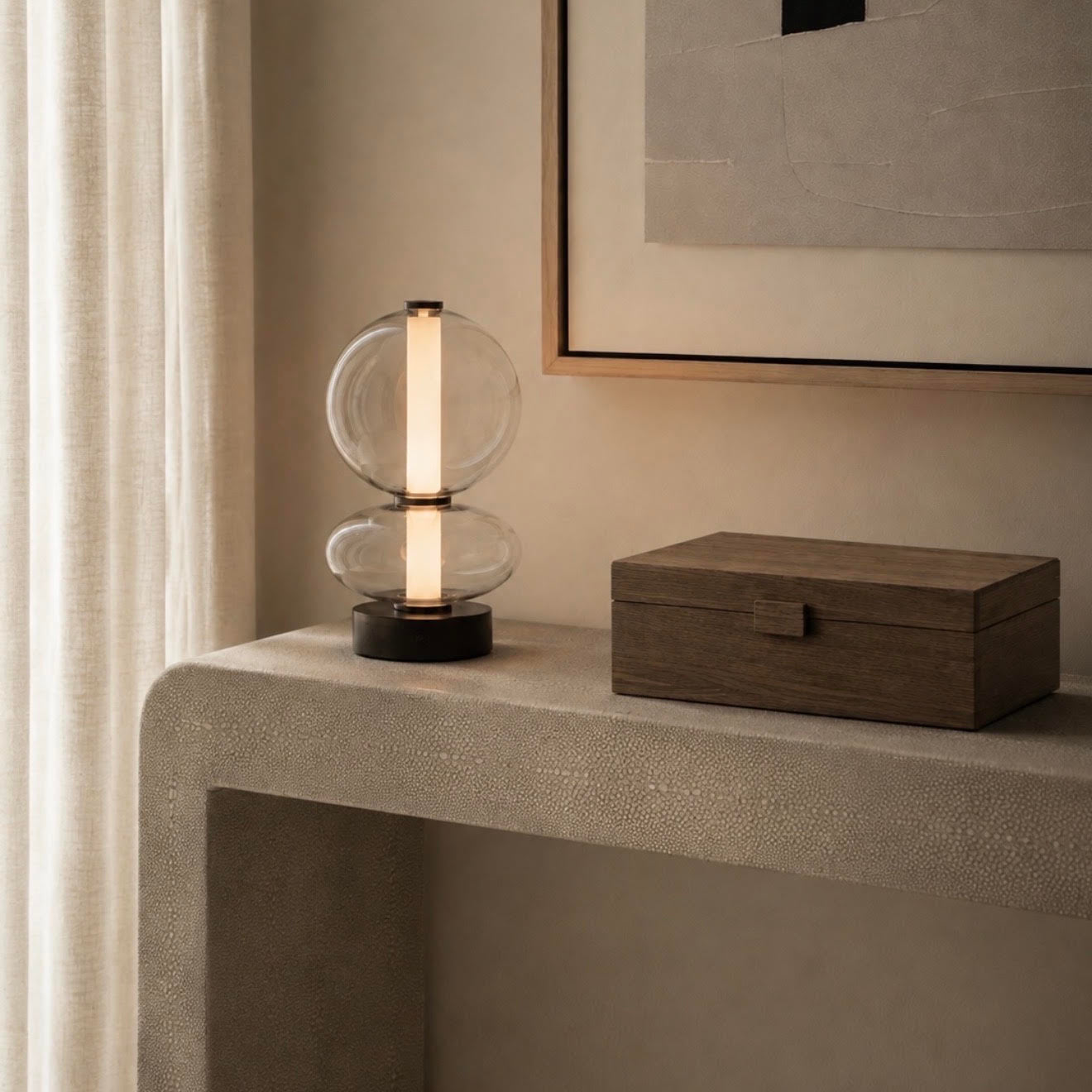 A modern table lamp with a clear glass double-sphere design and a vertical illuminated light bar, placed on a textured stone console table next to a dark wood box in a minimalist interior setting.