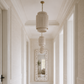 A long hallway with white paneled walls and ceiling, a woven beige runner rug, and a hanging cylindrical pendant light centered in the distance.
