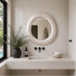 A round textured white mirror with a hammered finish, mounted on a light beige tiled bathroom wall above a floating white vanity with matte black fixtures.