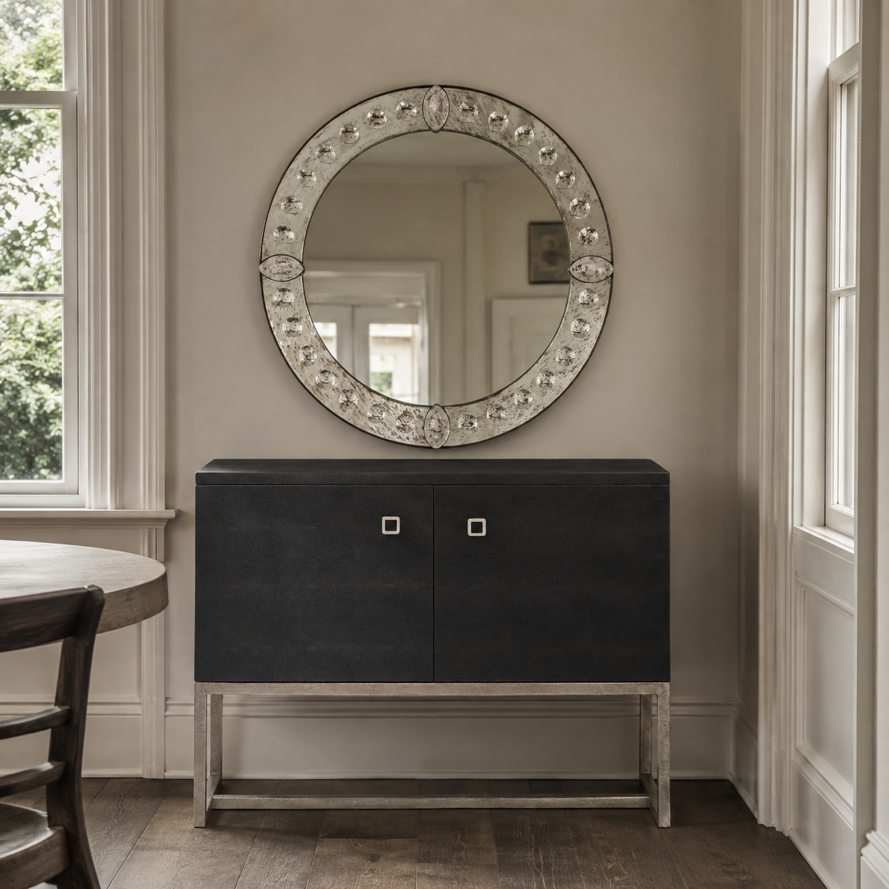 A round decorative wall mirror with an ornate silver-toned frame sits above a dark wood cabinet in a neutral-toned room with white trim and wood floors.