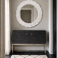 A modern entryway scene featuring a large, wavy-edged white oval mirror mounted on a gray wall above a dark charcoal gray console cabinet with a gold metal frame. A black and white patterned rug is on the floor, and a window with beige curtains is visible on the left.