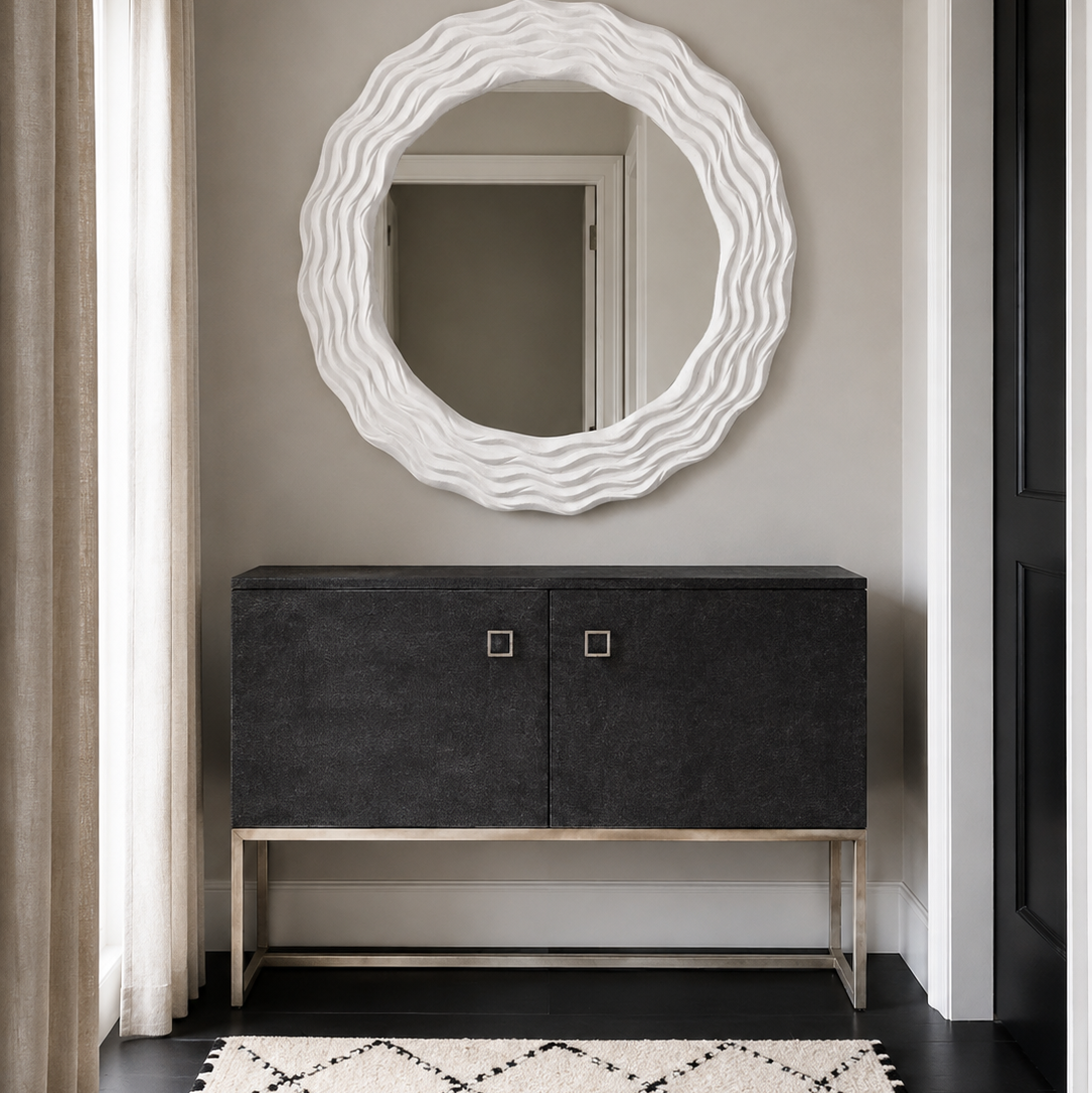 A modern entryway scene featuring a large, wavy-edged white oval mirror mounted on a gray wall above a dark charcoal gray console cabinet with a gold metal frame. A black and white patterned rug is on the floor, and a window with beige curtains is visible on the left.