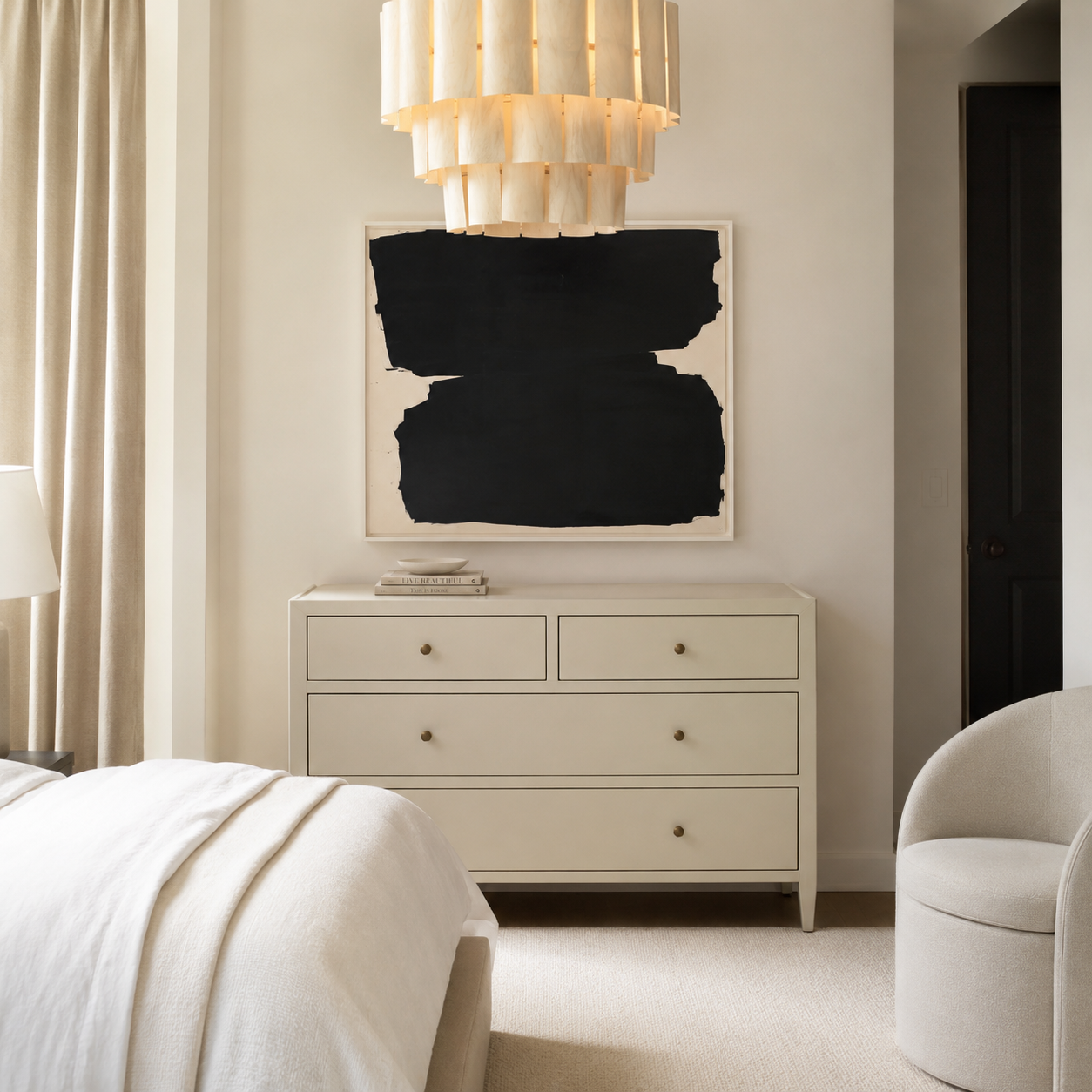 A styled bedroom interior featuring a large abstract black and white canvas print hanging on a beige wall above a cream-colored dresser. A modern tiered shell chandelier hangs from the ceiling, and a neutral-toned armchair is visible in the foreground.