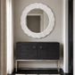 A modern entryway scene featuring a large, wavy-edged white oval mirror mounted on a gray wall above a dark charcoal gray console cabinet with a gold metal frame. A black and white patterned rug is on the floor, and a window with beige curtains is visible on the left.
