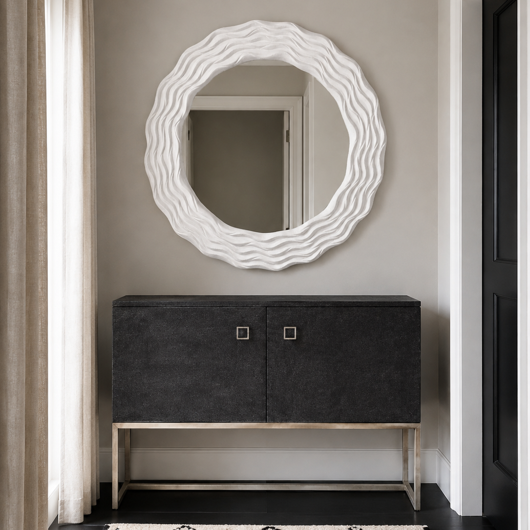 A modern entryway scene featuring a large, wavy-edged white oval mirror mounted on a gray wall above a dark charcoal gray console cabinet with a gold metal frame. A black and white patterned rug is on the floor, and a window with beige curtains is visible on the left.