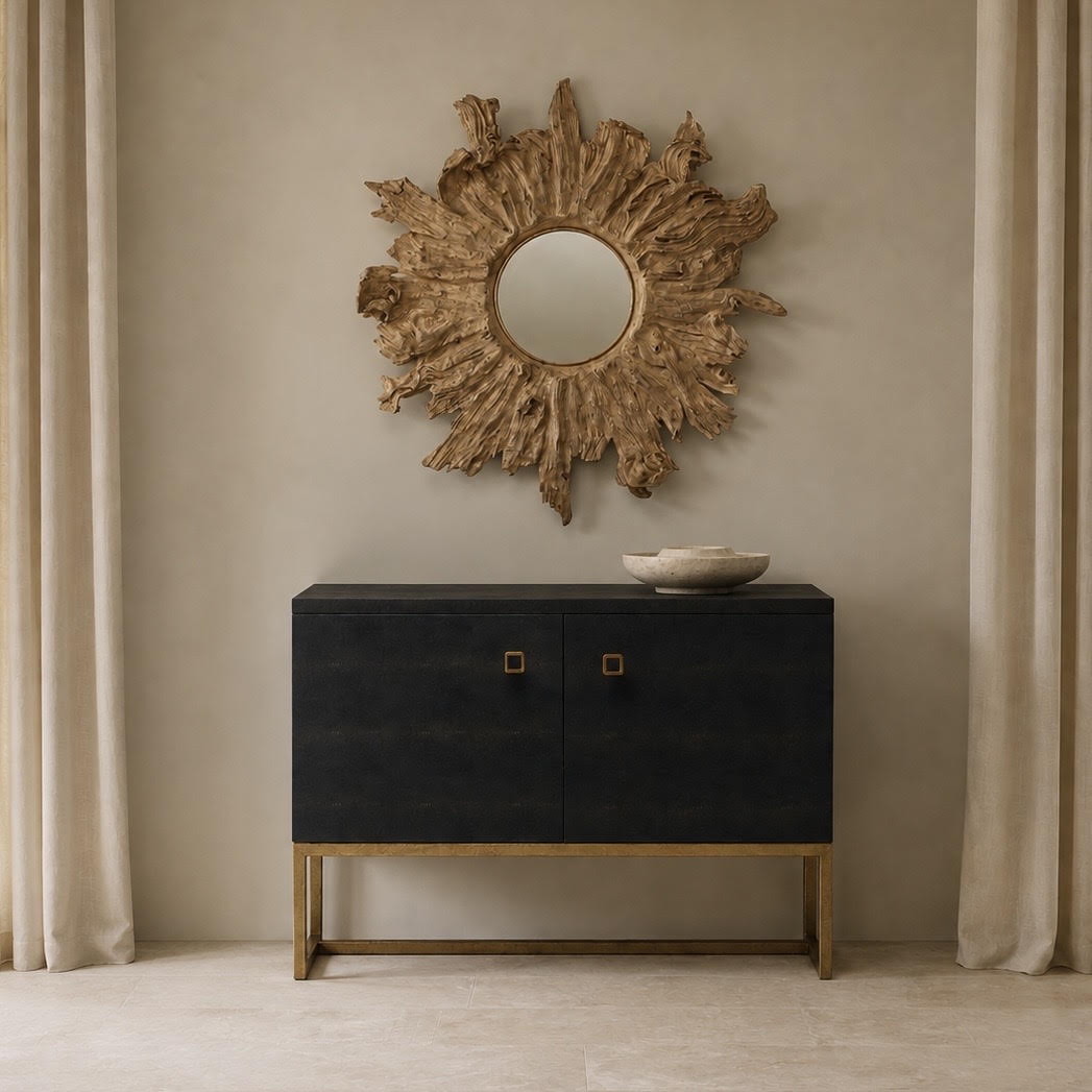 A modern black wood credenza with gold legs and square gold handles, styled in a room setting under a rustic sunburst mirror.