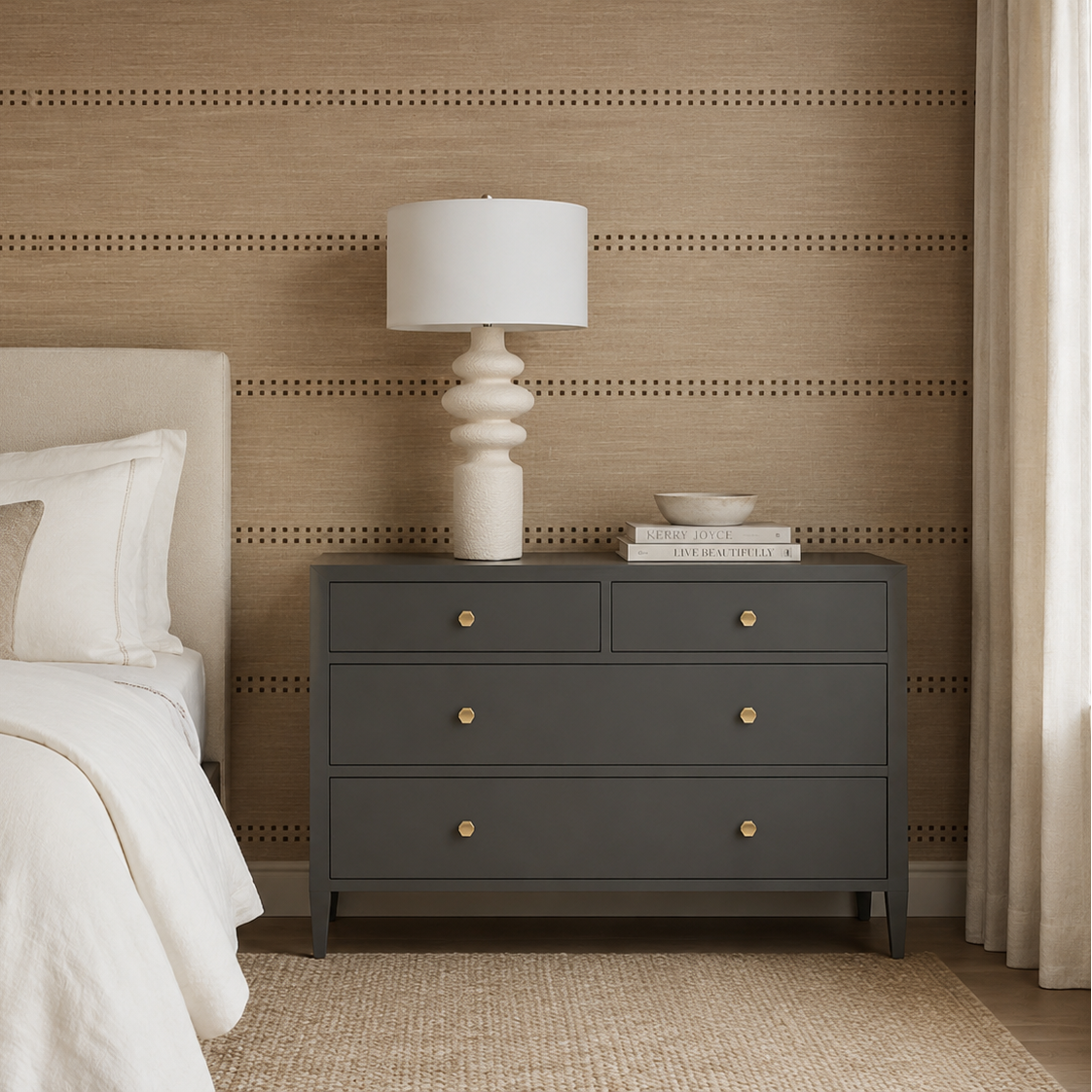 A dark gray three-drawer nightstand in a styled bedroom, featuring brass knobs, a white ceramic sculptural table lamp, and a small stack of books on top.