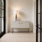 A modern light beige sideboard cabinet with two doors, square metal knobs, and a slim metal frame, styled in a minimalist entryway with a lit table lamp on top, light wood floors, and black-framed glass doors on both sides.