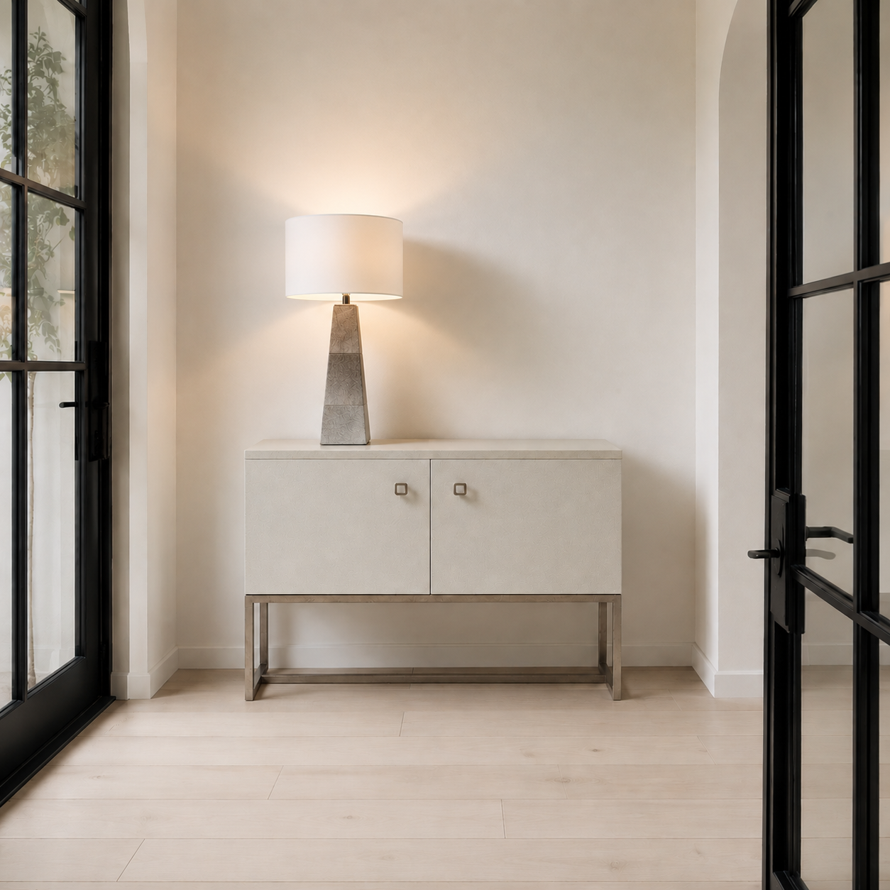 A modern light beige sideboard cabinet with two doors, square metal knobs, and a slim metal frame, styled in a minimalist entryway with a lit table lamp on top, light wood floors, and black-framed glass doors on both sides.
