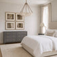 A styled bedroom interior featuring a gray six-drawer dresser with brass knobs, four framed black-and-white architectural prints, a white bed, a cream pendant light, and a window with sheer curtains.
