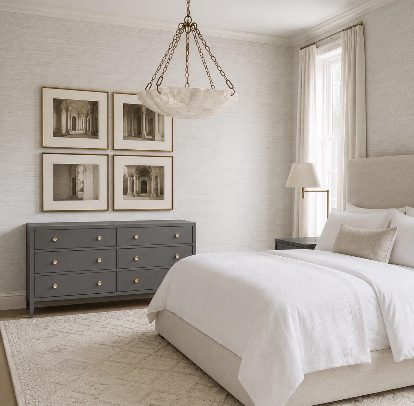 A styled bedroom interior featuring a gray six-drawer dresser with brass knobs, four framed black-and-white architectural prints, a white bed, a cream pendant light, and a window with sheer curtains.