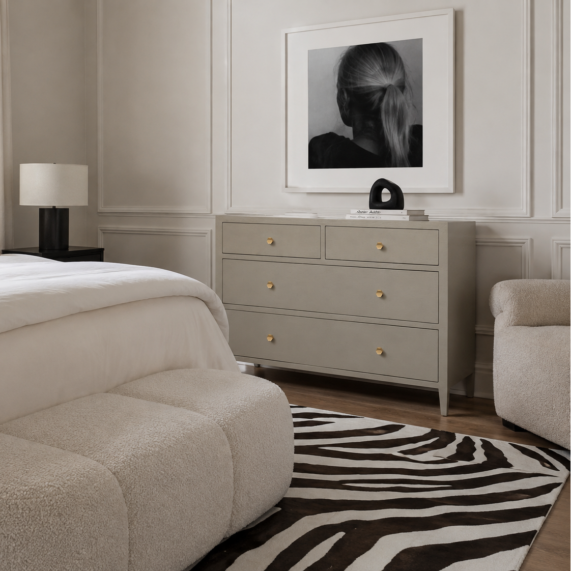A styled bedroom interior featuring a light gray three-drawer dresser with small gold knobs, a zebra-print rug, a tufted cream bench, and a black-and-white framed portrait on the wall.