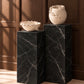 Two cream-colored decorative ceramic vessels, a bowl and a vase with a sculptural chain-like handle, displayed on black marble pedestals against a dark wood paneled wall.