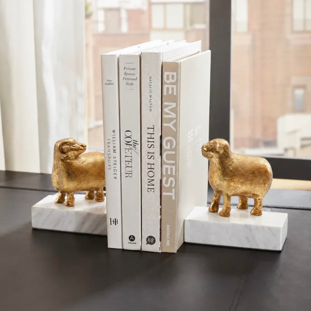 A pair of gold-finished sheep bookends on white marble bases holding a set of four books on a dark tabletop in front of a window.