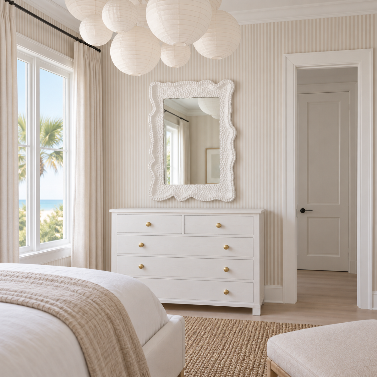 A white three-drawer dresser with small gold knobs in a bright, coastal-style bedroom. A white textured mirror with a scalloped frame hangs above the dresser, and a window on the left shows a view of palm trees and the ocean.