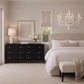 A styled bedroom interior featuring a black six-drawer dresser with small gold knobs, a white table lamp, a vase of white flowers, a beige upholstered bed, a white chandelier, and a beige bench with a throw blanket.