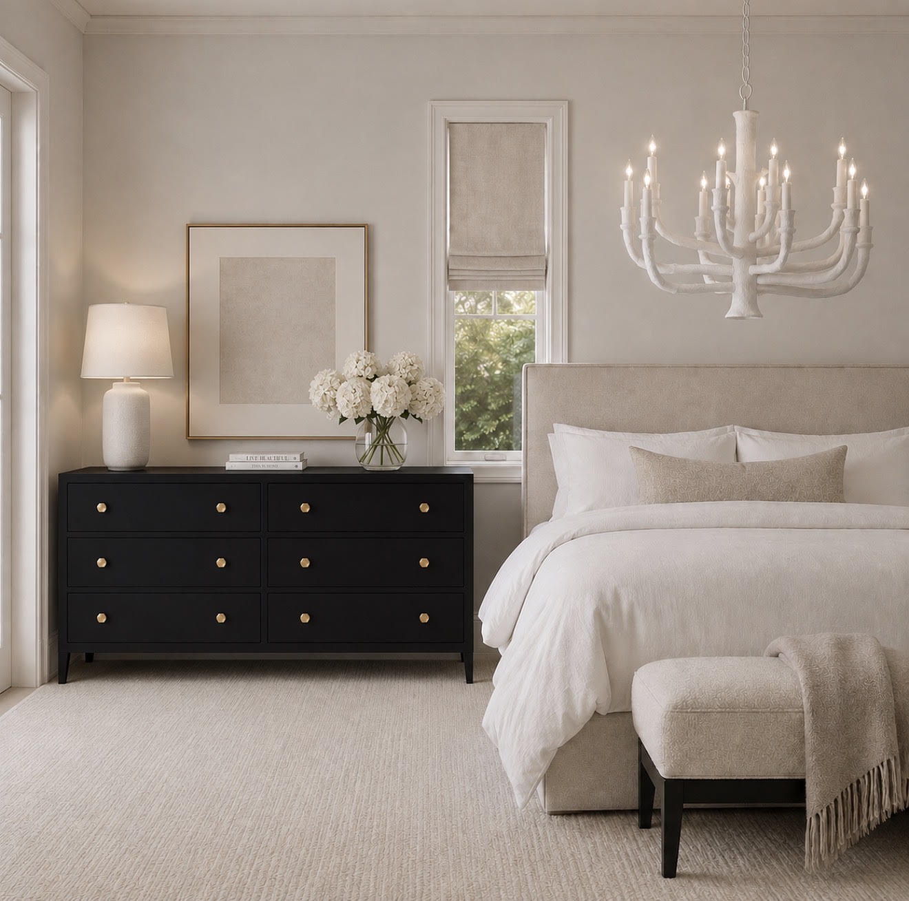A styled bedroom interior featuring a black six-drawer dresser with small gold knobs, a white table lamp, a vase of white flowers, a beige upholstered bed, a white chandelier, and a beige bench with a throw blanket.