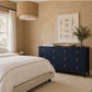 A navy blue wooden dresser with gold knobs in a styled bedroom setting, featuring a white bed, textured beige walls, a woven pendant light, and a framed art print.