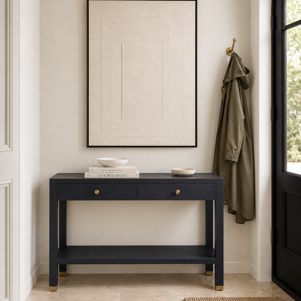 A navy blue console table with two drawers and brass hardware stands against a light beige wall in a styled entryway. A framed abstract print hangs above, a green coat is hanging on a gold hook to the right, and a window is visible on the right edge.