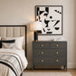 A dark gray three-drawer nightstand with gold knobs in a styled bedroom setting, next to a bed with neutral bedding and a lit table lamp. A framed black and white abstract print hangs on the beige wall above the nightstand.