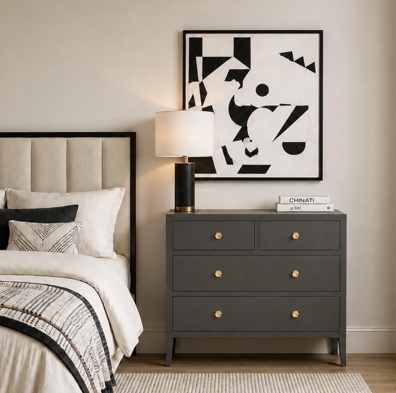 A dark gray three-drawer nightstand with gold knobs in a styled bedroom setting, next to a bed with neutral bedding and a lit table lamp. A framed black and white abstract print hangs on the beige wall above the nightstand.