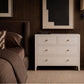 A white three-drawer dresser with small round gold knobs stands beside a bed in a dark wood bedroom setting.