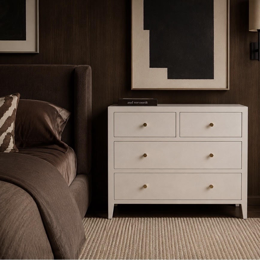 A white three-drawer dresser with small round gold knobs stands beside a bed in a dark wood bedroom setting.