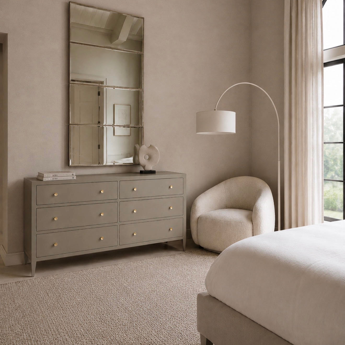 A styled bedroom interior featuring a light gray dresser with gold knobs and a large rectangular mirror, next to a cream upholstered accent chair and a white floor lamp. A bed with white bedding is partially visible in the foreground, with beige curtains and a window in the background.