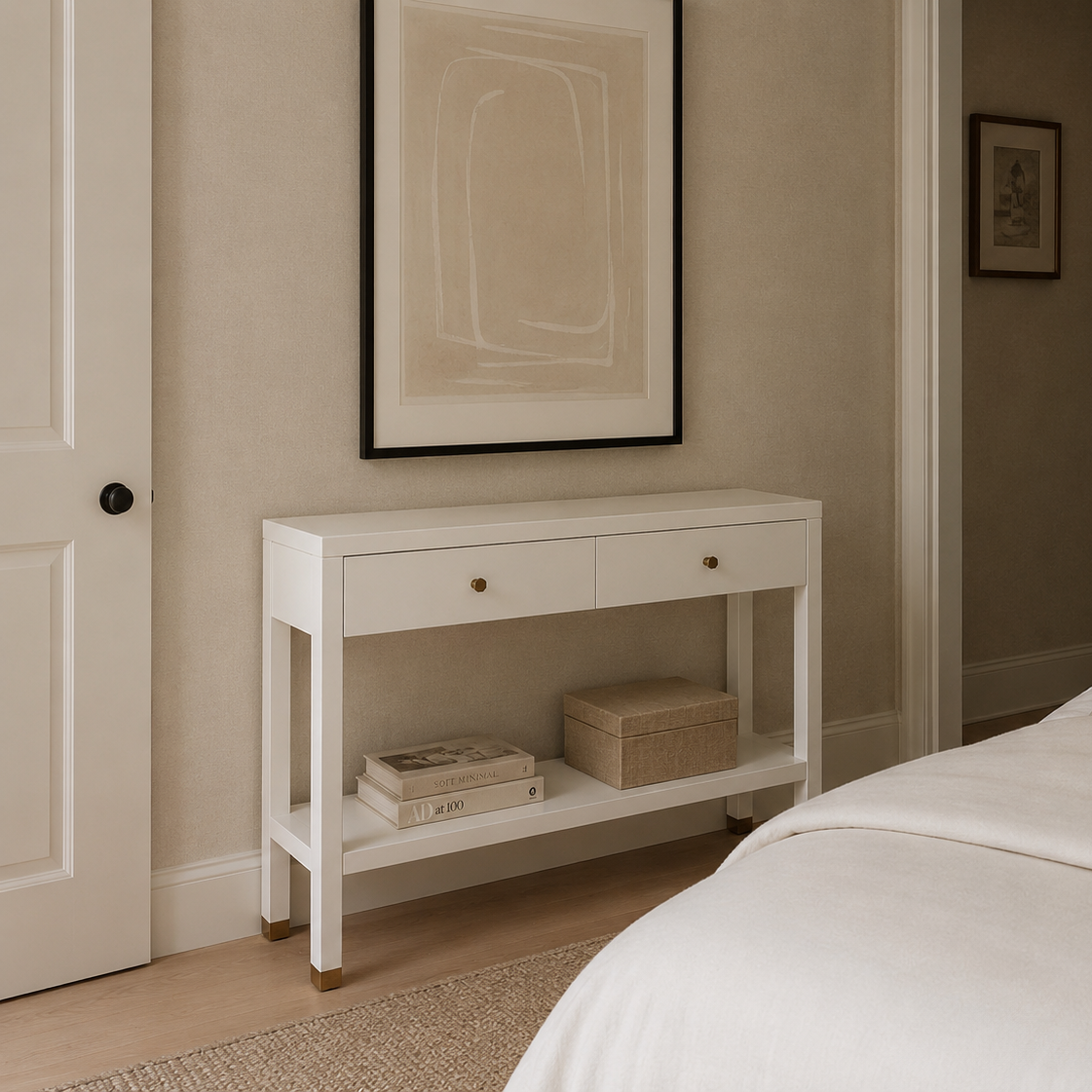 A white two-drawer console table with open shelving stands beside a bed in a softly styled bedroom. The table has small round gold-toned knobs and holds a book and a woven box on the lower shelf.