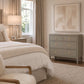 A gray three-drawer dresser with small gold knobs in a styled bedroom setting, next to a bed with neutral bedding and a window with beige curtains.