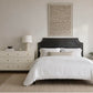 A styled bedroom scene featuring a bed dressed in white linens with a dark wooden headboard, a cream dresser with a lamp, and a large framed abstract artwork on the wall.