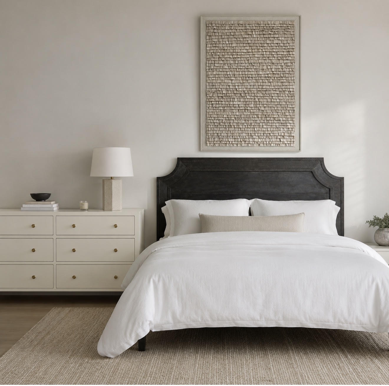 A styled bedroom scene featuring a bed dressed in white linens with a dark wooden headboard, a cream dresser with a lamp, and a large framed abstract artwork on the wall.