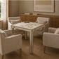 A square wooden table with a light beige top and cream legs, surrounded by four matching upholstered armchairs in a warm neutral living room setting.