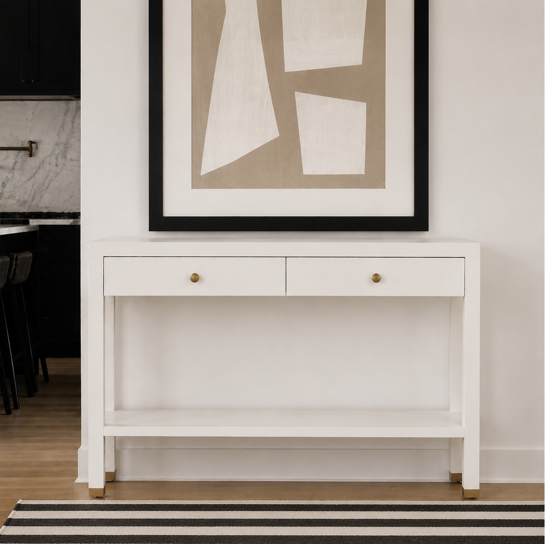 A white console table with two drawers and brass knobs, styled in a modern home interior with a black-framed abstract print on the wall above it.