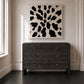 A dark charcoal gray wooden dresser with brass handles, positioned against a white paneled wall below a black and white abstract canvas print.