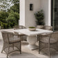 A 5-piece outdoor dining set featuring a square white stone table with a round pedestal base and four woven wicker chairs, set on a patio with a bowl of greenery on top.