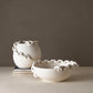 Two matte white ceramic decorative pieces, a round vase and a shallow bowl, both featuring sculptural chain-like handles around the rims, displayed on a neutral beige surface against a taupe background.